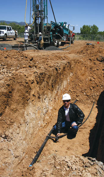 While one of his drilling rigs (background) bores another of the 112 holes at Holmes Middle School, Mike Everett of 
Rocky Mountain Geo-thermal shows where a pipe goes down one of the existing 300-foot-deep holes and connects
to a system supply line about 6 feet below the field surface. 
Westside Pioneer photo