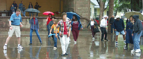 A local line-dancing group directed by Coleen Weis (rear, 
with red umbrella) splashes with eager amateurs to the 
rhythm of the music and the rain at the annual West 
Center/Organization of Westside Neighbors Block Party/Picnic at Bancroft Park June 27.